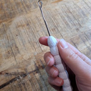 Puede incluir: Un ganchillo plateado con un lazo en el extremo, sostenido en una mano. El mango es de color beige claro y tiene un diseño texturizado y acanalado. El ganchillo está diseñado para manualidades y trabajos textiles.