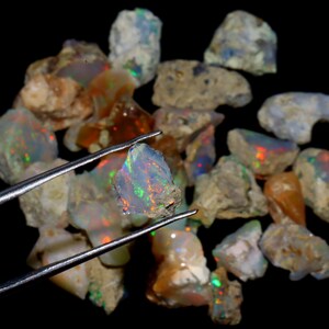 May include: A collection of rough opal stones, some with iridescent colors, in various sizes and shapes. One opal stone is held by tweezers, showcasing its rainbow-like sheen.