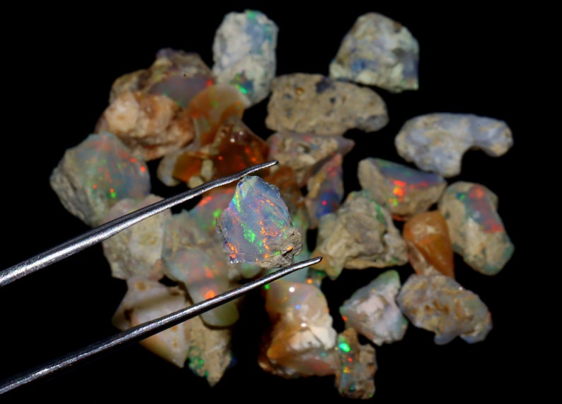May include: A collection of rough opal stones, some with iridescent colors, in various sizes and shapes. One opal stone is held by tweezers, showcasing its rainbow-like sheen.