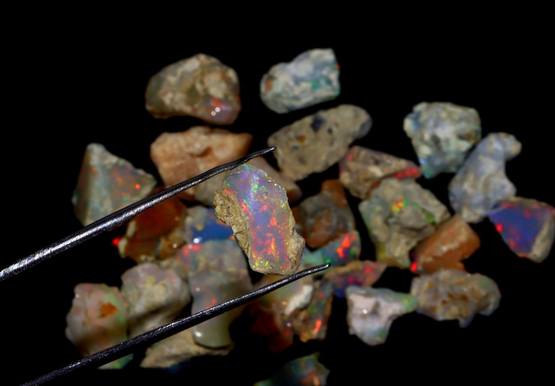 May include: A collection of rough opal stones, some with iridescent colors, are displayed on a black surface. One stone is held by tweezers, showcasing its rainbow-like sheen.