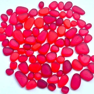 May include: A collection of numerous small, smooth, rounded pieces of red sea glass scattered across a plain white surface. The pieces vary in size and shape, displaying translucent and opaque shades of bright red, deep crimson, and pinkish tones.