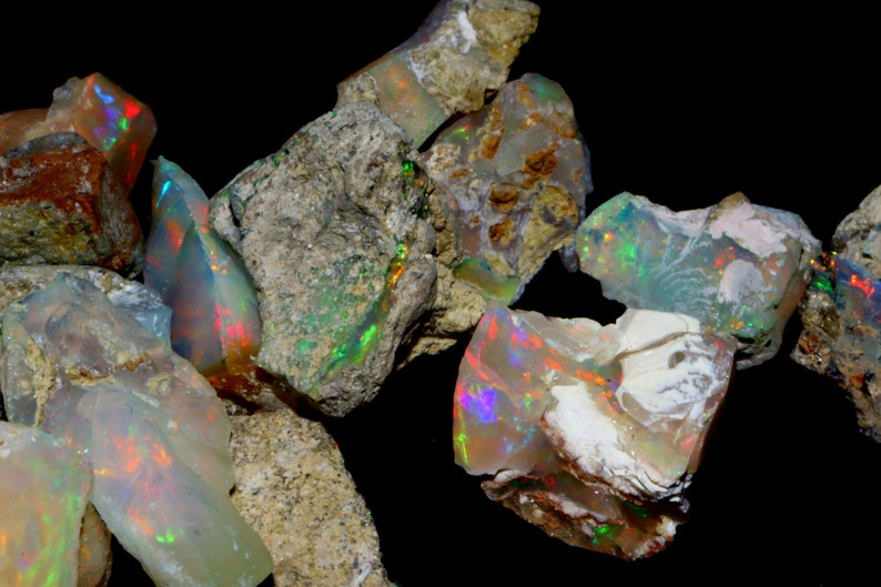 May include: A collection of rough opal stones with iridescent colors, including green, blue, red, and orange. The stones are embedded in a gray, brown, and white matrix.