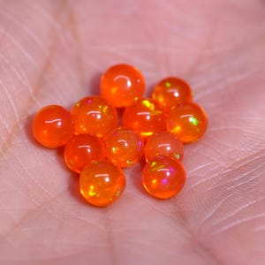 Puede incluir: Un grupo de pequeñas cuentas de ópalo redondas y naranjas con un brillo iridiscente. Las cuentas están dispuestas en un grupo suelto en una mano.