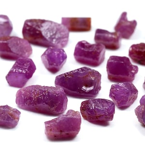 Puede incluir: Una colección de pequeños cristales de rubí rosa en bruto. Los cristales son de diversas formas y tamaños, algunos tienen una forma más puntiaguda y otros son más redondeados.