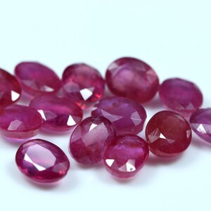 Puede incluir: Un grupo de piedras preciosas de rubí de forma ovalada y facetadas en varios tonos de rosa y rojo. Las piedras preciosas están dispuestas en un montón suelto sobre un fondo blanco.