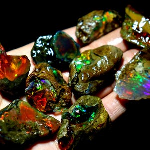Puede incluir: Una colección de piedras de ópalo en bruto con colores iridiscentes, incluyendo verde, azul, rojo y naranja. Las piedras son de varias formas y tamaños y están dispuestas sobre un fondo oscuro.