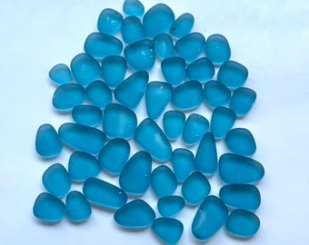 May include: A collection of numerous small, smooth, translucent blue glass pieces with rounded, irregular shapes. These individual glass fragments feature a matte, frosted texture and vary in size, creating a uniform bright blue color palette.