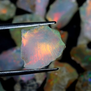 Op de afbeelding: Een close-up van een kleine, iriserende opaal edelsteen met een regenboog aan kleuren. De opaal wordt vastgehouden door een pincet en is omringd door andere opalen.