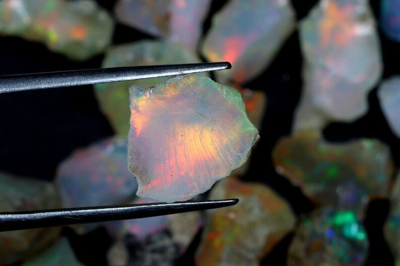 Op de afbeelding: Een close-up van een kleine, iriserende opaal edelsteen met een regenboog aan kleuren. De opaal wordt vastgehouden door een pincet en is omringd door andere opalen.