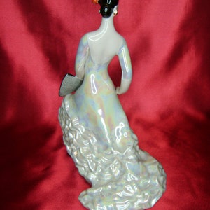 Puede incluir: Una figurita de porcelana de una mujer con un vestido fluido e iridiscente. Lleva un abanico en la mano izquierda y la derecha levantada. La figurita est&aacute; sobre una superficie roja.