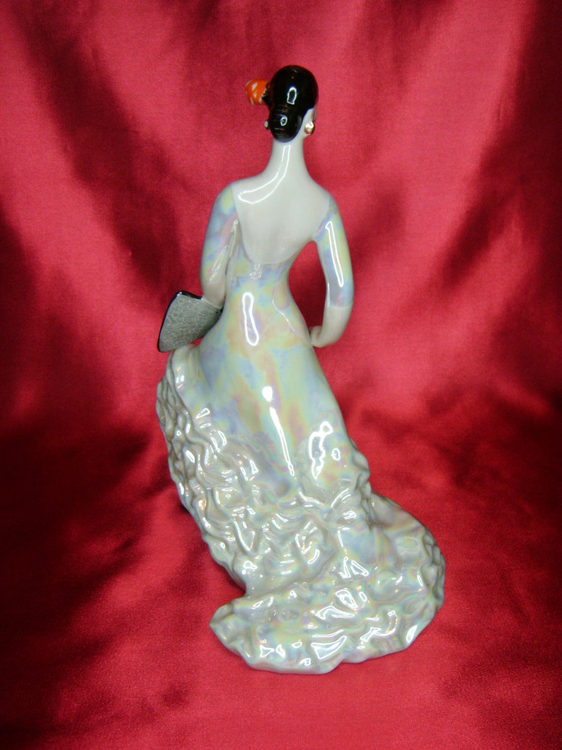 Puede incluir: Una figurita de porcelana de una mujer con un vestido fluido e iridiscente. Lleva un abanico en la mano izquierda y la derecha levantada. La figurita est&aacute; sobre una superficie roja.