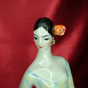 Puede incluir: Una figurita de porcelana de una mujer con cabello negro y una flor roja en el cabello. Lleva un vestido blanco con un patr&oacute;n azul claro y rosa.