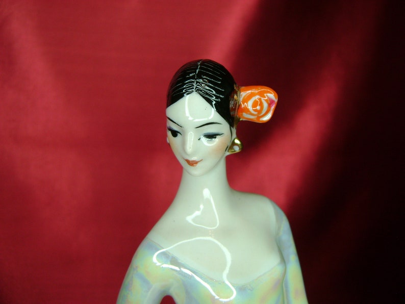 Puede incluir: Una figurita de porcelana de una mujer con cabello negro y una flor roja en el cabello. Lleva un vestido blanco con un patr&oacute;n azul claro y rosa.