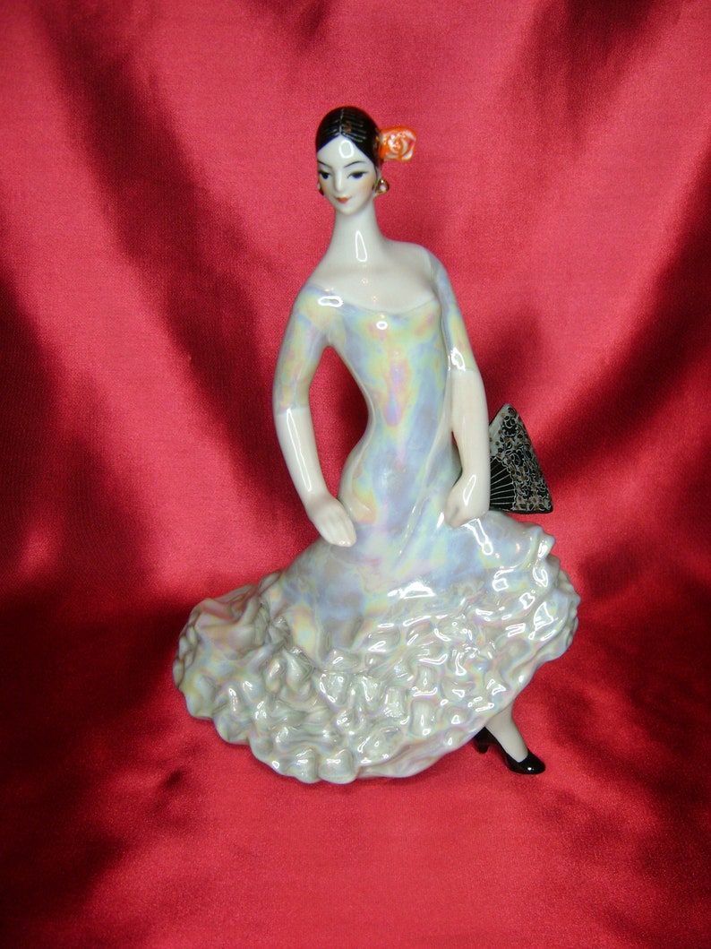 Puede incluir: Una figurita de porcelana de una mujer con un vestido blanco e iridiscente con una falda con volantes. Lleva un abanico negro y una flor roja en el pelo.
