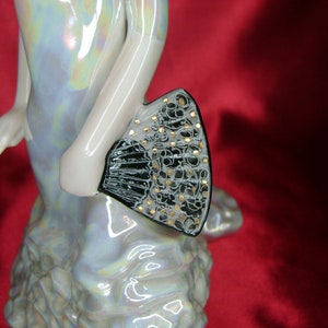 Puede incluir: Una figurita de porcelana de una mujer con un vestido blanco y un abanico negro y dorado. El vestido tiene un acabado iridiscente nacarado.