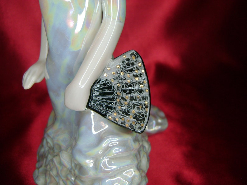 Puede incluir: Una figurita de porcelana de una mujer con un vestido blanco y un abanico negro y dorado. El vestido tiene un acabado iridiscente nacarado.