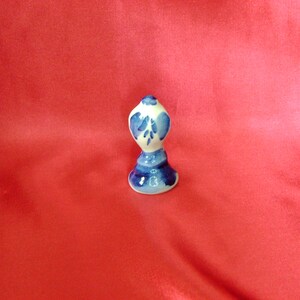 Puede incluir: Un peque&ntilde;o tap&oacute;n de botella de cer&aacute;mica blanca con un dise&ntilde;o floral azul y blanco. El tap&oacute;n tiene forma de campana con la parte superior redondeada.