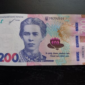 May include: A Ukrainian 200-hryvnia banknote. The front features a portrait of a woman, with the text "НАЦІОНАЛЬНИЙ БАНК УКРАЇНИ" at the top. The number "200" is prominently displayed in blue and pink.