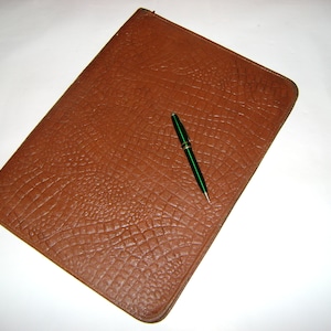 Puede incluir: Una carpeta de cuero marrón con una superficie texturizada y un bolígrafo verde encima.