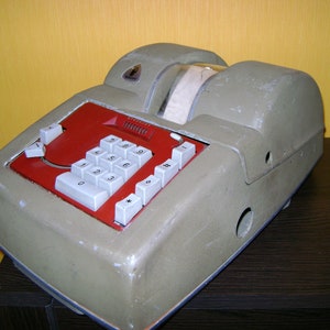 Op de afbeelding: Een antieke kassa in olijfgroen met een rood toetsenbord en witte knoppen. Het heeft een papierrol dispenser. De machine staat op een donkerbruin oppervlak. Het toetsenbord heeft een numerieke indeling.