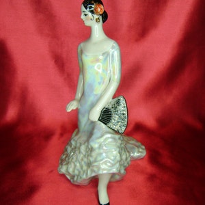 Puede incluir: Una figurita de porcelana de una mujer con un vestido blanco e iridiscente, sosteniendo un abanico negro y plateado. Tiene el pelo negro y una flor roja en el pelo.