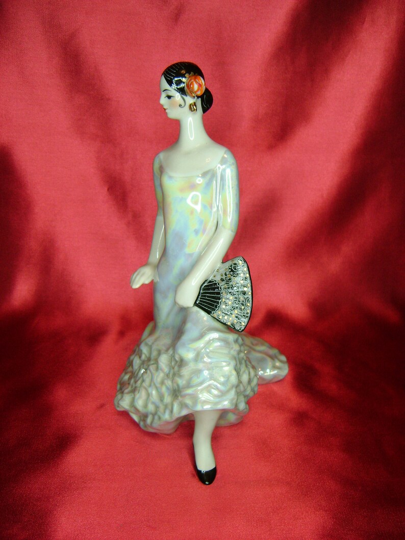 Puede incluir: Una figurita de porcelana de una mujer con un vestido blanco e iridiscente, sosteniendo un abanico negro y plateado. Tiene el pelo negro y una flor roja en el pelo.