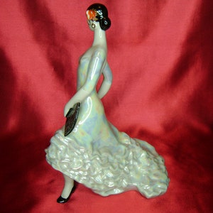 Puede incluir: Una figurita de porcelana de una mujer con un vestido azul iridiscente con volantes. Lleva un abanico en la mano izquierda y una flor roja en el pelo.