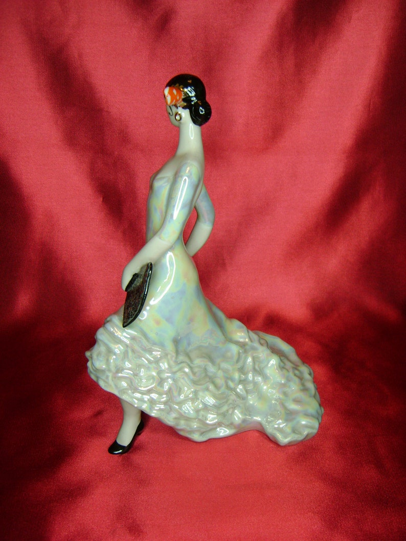 Puede incluir: Una figurita de porcelana de una mujer con un vestido azul iridiscente con volantes. Lleva un abanico en la mano izquierda y una flor roja en el pelo.