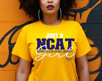 May include: A yellow t-shirt with the text "JUST A NCAT HISTORICALLY BLACK COLLEGE & UNIVERSITY" in blue and white. The word "Aggie" is written in script. The shirt is worn against an orange and black painted wall.