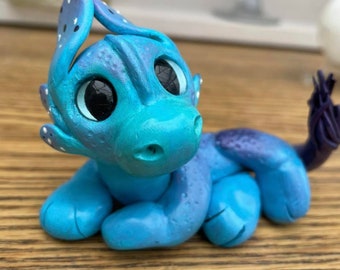 Op de afbeelding: Een klein, blauw en paars drakenbeeldje met grote ogen en een gevlekte kop. De draak ligt op zijn buik met zijn staart opgerold.