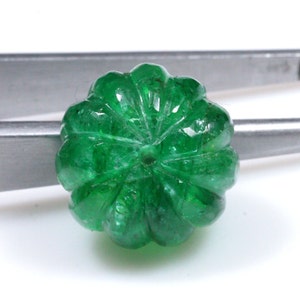 May include: A deep green emerald gemstone carved into a round, fluted shape. The gemstone has a hole drilled through the center.