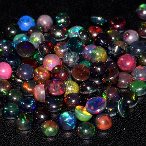 May include: A collection of round, iridescent black opals in various shades of green, red, blue, and purple. The opals are arranged in a loose pile on a black surface.