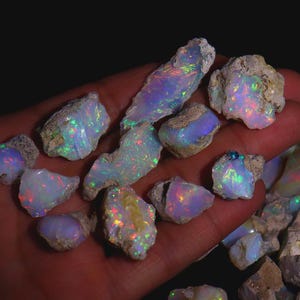 Può includere: Una collezione di opali grezzi, che mostra uno spettro di colori tra cui blu, verdi e rossi. Gli opali hanno una consistenza ruvida e naturale e sono tenuti in una mano su uno sfondo scuro. Queste pietre preziose sono ideali per la creazione di gioielli.