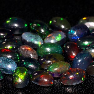 May include: A collection of oval-shaped black opals with iridescent green, blue, and red flashes. The opals are arranged in a loose pile on a black background.