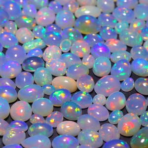 Puede incluir: Una colección de cabujones de ópalo iridiscente en varios tamaños y formas. Los ópalos son de color blanco lechoso con destellos de colores arcoíris.