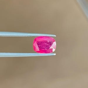 May include: A vibrant, cushion-cut pinkish-red gemstone held securely between the tips of a pair of silver-toned metal tweezers. The stone features a faceted surface that reflects light, showcasing a deep, saturated color and a clear, polished appearance.