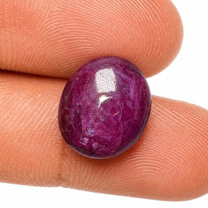 Op de afbeelding: Een diep magenta-kleurige ovale edelsteen met een glad, gepolijst cabochon oppervlak. De steen vertoont een doorschijnende kwaliteit met interne insluitingen en een reflecterende highlight op de afgeronde bovenkant, met een rijke kleur.