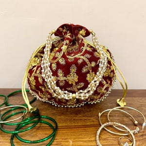 May include: A deep red velvet drawstring pouch featuring intricate gold embroidery and sequin floral patterns, accented with a multi-strand pearl handle. The pouch is surrounded by several green bangles, gold-toned bracelets, and a small ring.