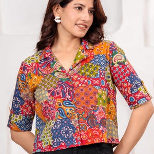 May include: A button-down shirt featuring a vibrant patchwork pattern with various paisley, floral, and geometric motifs in shades of red, orange, blue, green, and yellow. The shirt has a collared neckline, short sleeves with cuffs, and a cropped hem.