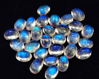 May include: A collection of oval-shaped moonstone cabochons, with a milky white color and iridescent blue flashes. The stones are arranged in a loose pile on a black background.