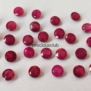 May include: A collection of small, round, faceted red rubies scattered across a plain surface. Each gemstone displays a vibrant crimson hue with multiple cut facets that reflect light. The text "preciousclub" is printed in black in the center.