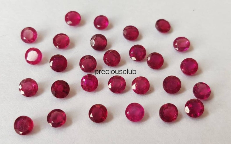 May include: A collection of small, round, faceted red rubies scattered across a plain surface. Each gemstone displays a vibrant crimson hue with multiple cut facets that reflect light. The text "preciousclub" is printed in black in the center.