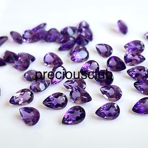 May include: A collection of pear-shaped amethyst gemstones in various sizes, displayed on a white surface. The gemstones are a deep purple colour and have a smooth, polished finish.