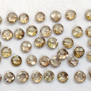 May include: A collection of 30 round, faceted, brown diamonds. The diamonds are loose and scattered on a white surface.