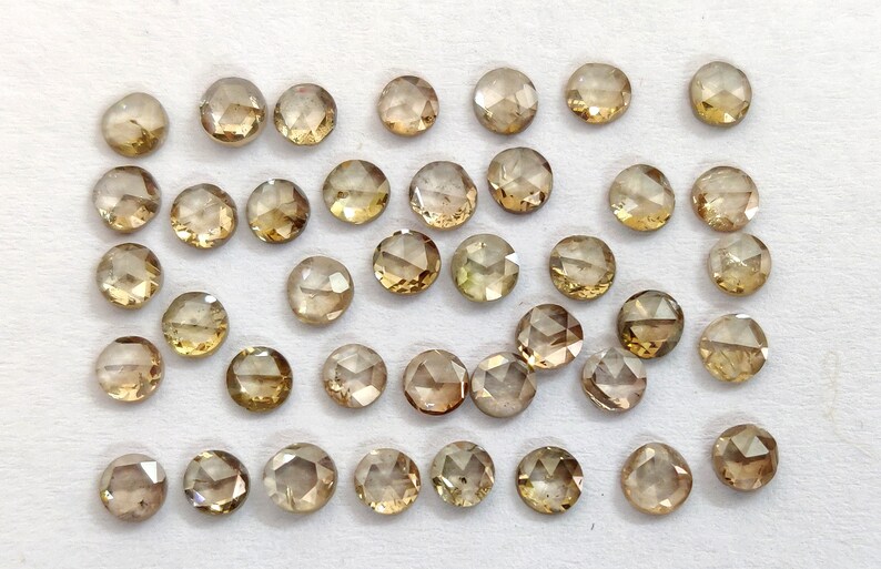 May include: A collection of 30 round, faceted, brown diamonds. The diamonds are loose and scattered on a white surface.