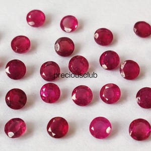 May include: A collection of twenty-six small, round, faceted red rubies arranged in a loose pattern. Each gemstone displays a vibrant crimson hue with multiple facets reflecting light. The text "preciousclub" is printed in black in the center.