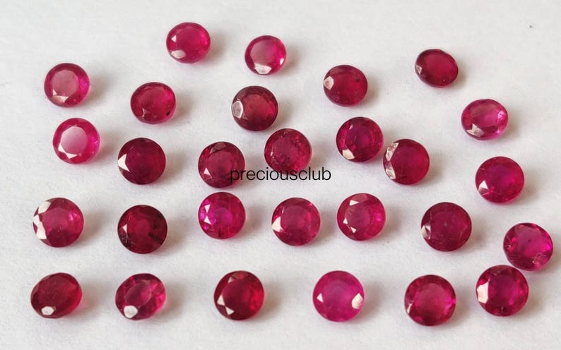 May include: A collection of twenty-six small, round, faceted red rubies arranged in a loose pattern. Each gemstone displays a vibrant crimson hue with multiple facets reflecting light. The text "preciousclub" is printed in black in the center.