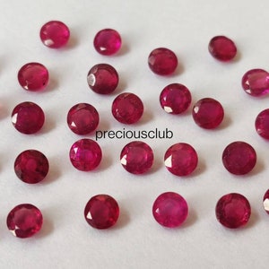 May include: A collection of small, round, faceted red rubies scattered across a plain surface. Each gemstone displays a brilliant cut with multiple reflective surfaces. The text "preciousclub" is printed in black in the center of the arrangement.
