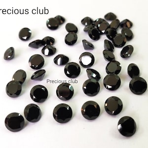 Puede incluir: Una colección de piedras preciosas redondas negras, probablemente diamantes, esparcidas sobre una superficie blanca. Las piedras preciosas son de varios tamaños y tienen un acabado brillante y pulido.