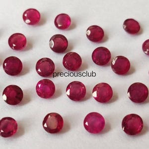 May include: A collection of small, round, faceted red rubies scattered across a plain surface. Each gemstone features a brilliant cut with multiple reflective surfaces. The text "preciousclub" is printed in black in the center of the arrangement.